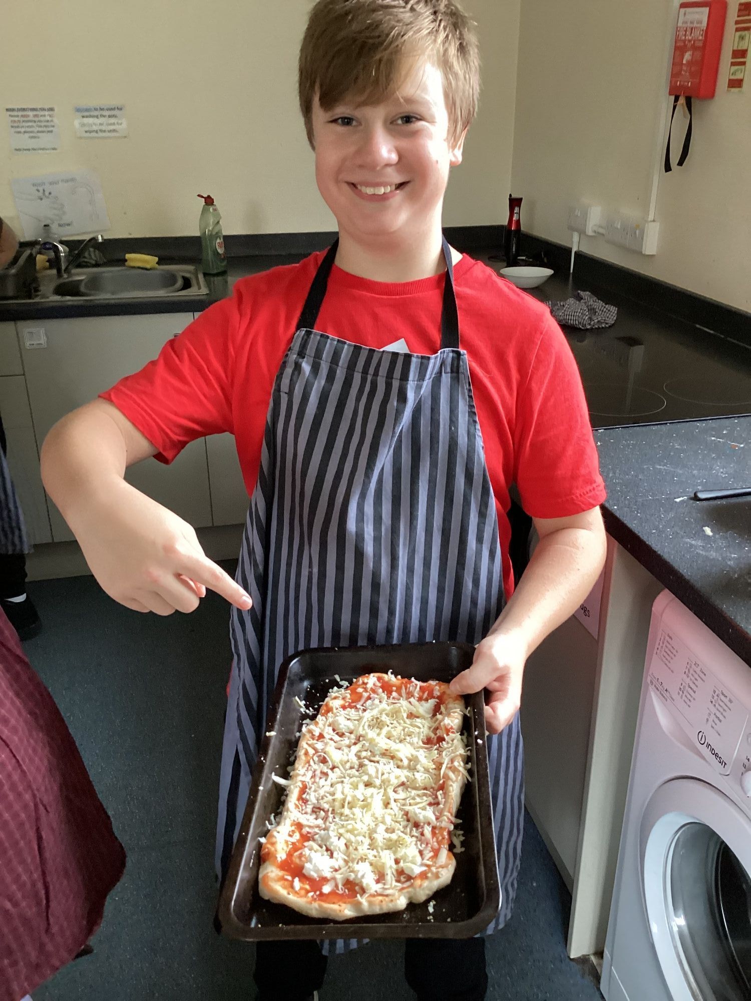 Boy in a red t-shirt and stripy apron pointing at the pizza he has made